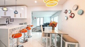 Eine moderne Küche mit weißen Schränken, einer Marmorarbeitsplatte und einer Bar mit orangefarbenen Hockern. Ein Esstisch mit Bänken und ein Kronleuchter sind ebenfalls vorhanden.