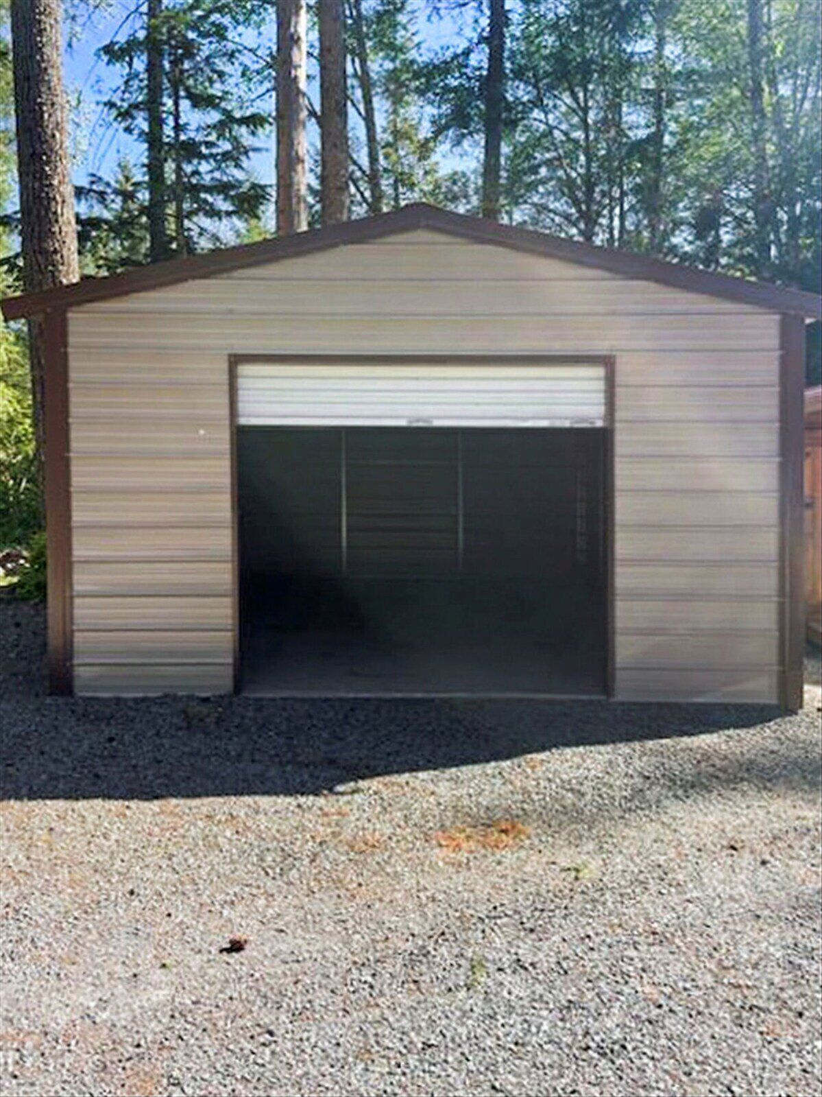 A single-story metal shed with a closed garage door, surrounded by gravel and trees.