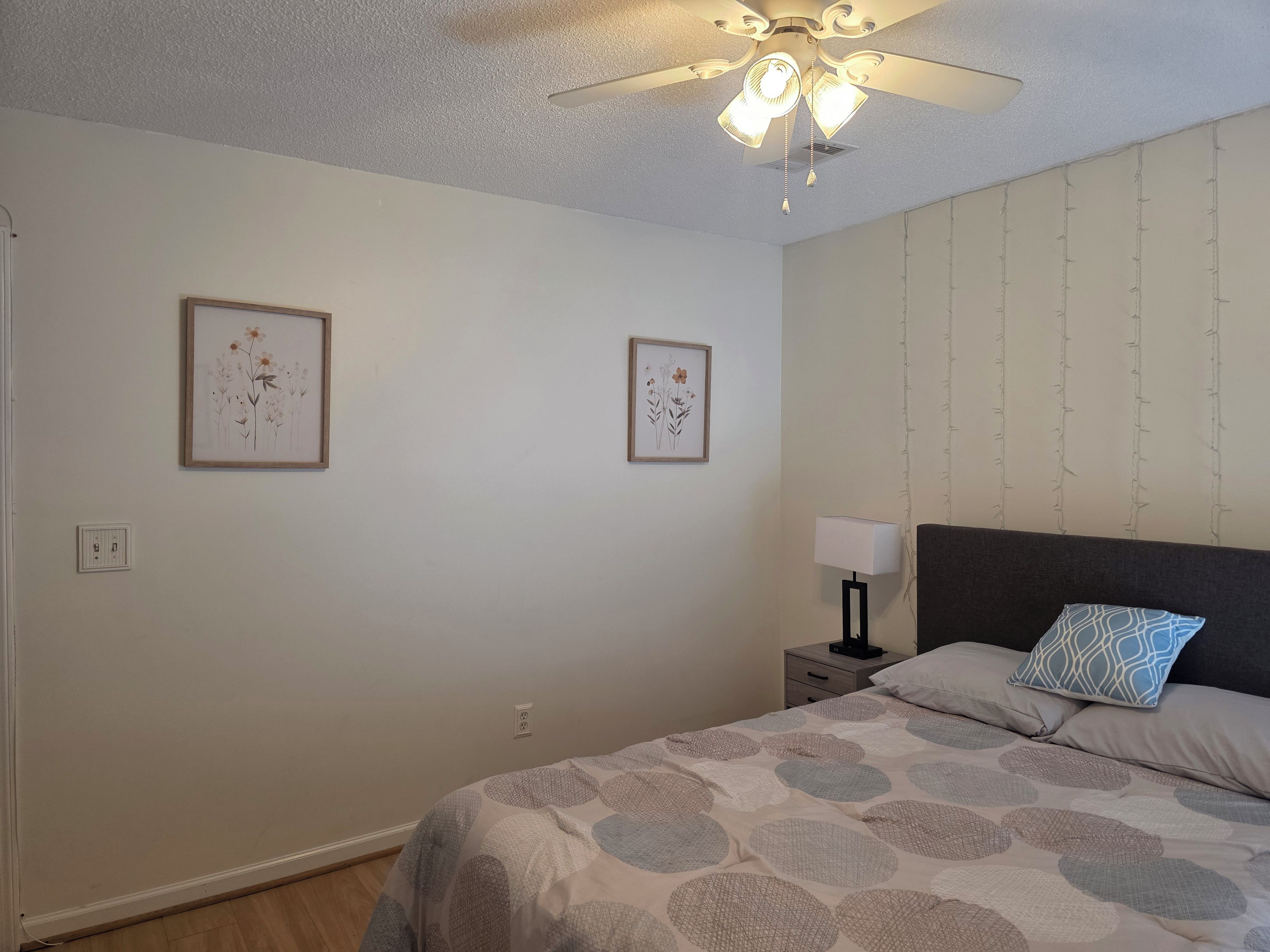 A bedroom with a bed, a nightstand, a ceiling fan, and two framed floral artworks on the wall.
