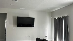 A living room with a flat-screen TV mounted on the wall, a black sofa, and a wooden floor. - Family Fun Getaway, Spacious, Cozy & Kid-Friendly (Ocala)