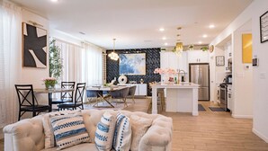 Uma cozinha moderna com ilha central, área de jantar com cadeiras e sala de estar com sofá e almofadas decorativas.