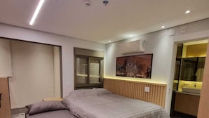 A modern bedroom with a bed, bedside tables, a window with blinds, a wall-mounted air conditioner, and a painting.