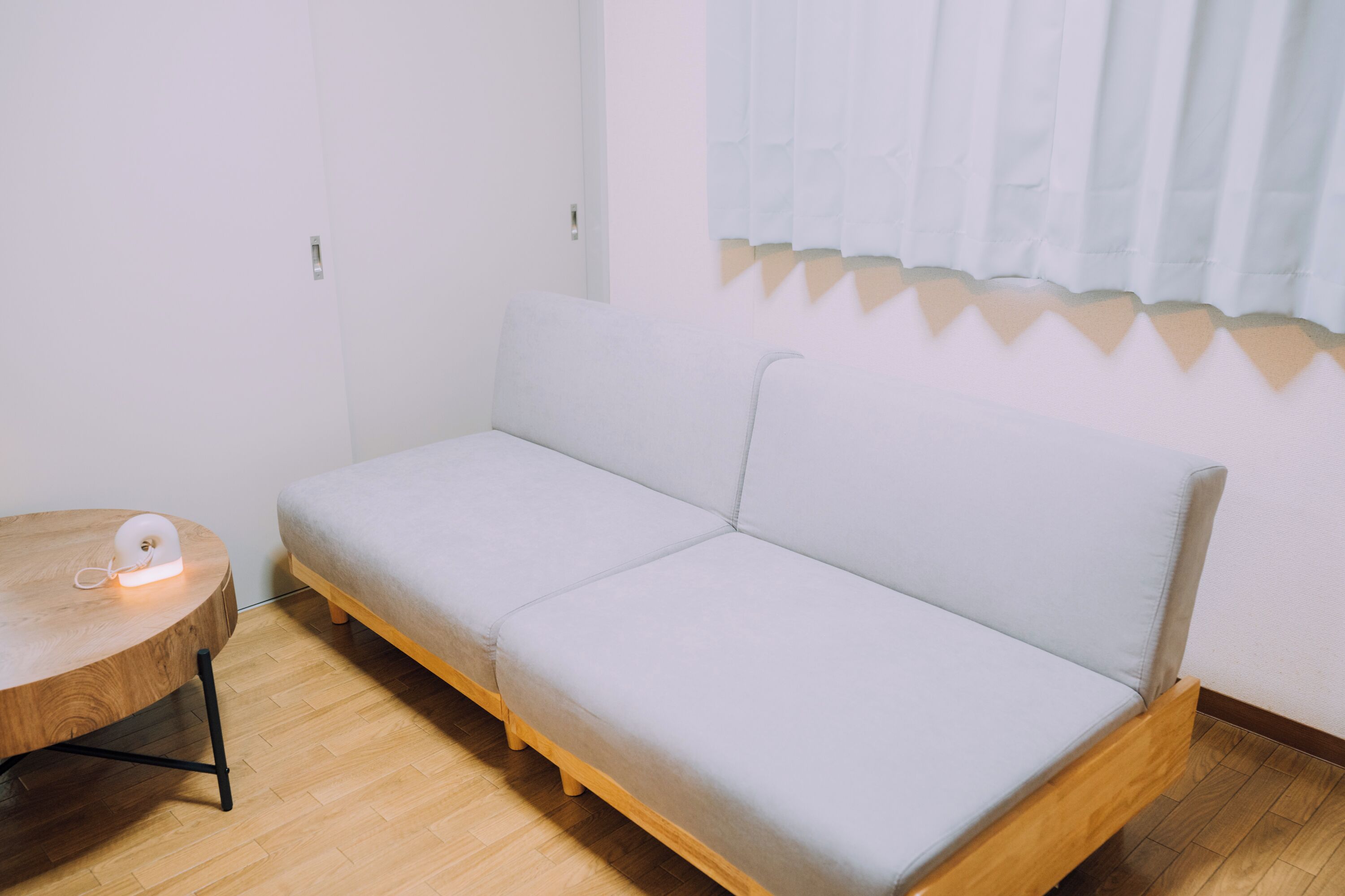 Ein Zimmer mit einem hellgrauen Sofa, einem runden Holztisch mit einer weißen Lampe und einem Fenster mit weißen Vorhängen.