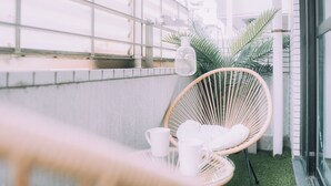 A wicker chair with a white cushion and a white bottle on a table, set against a white wall with a metal railing. - Showa Bloom · Near Shinjuku Station (Tokyo)