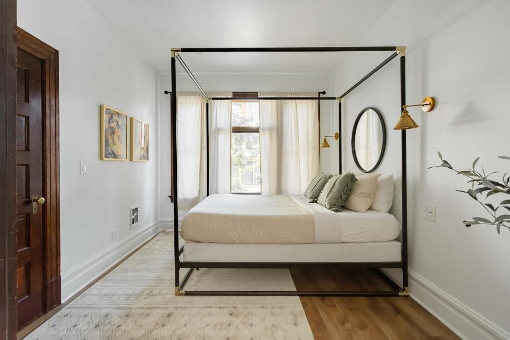 A four-poster bed in a room with a wooden floor, white walls, and a door on the left.