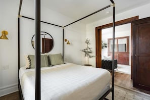 Ein Himmelbett mit weißem Überwurf, ein runder Spiegel darüber und ein kleiner Blumentopf daneben.