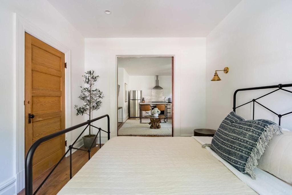 A bedroom with a bed, a wooden door, a potted plant, a kitchen in the background, and a small table with a lamp.