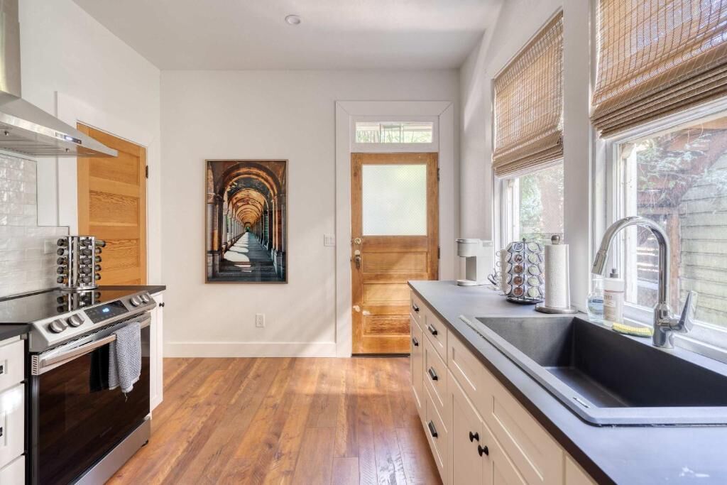 A kitchen with wooden flooring, a stove, a sink, and a door leading to another room.