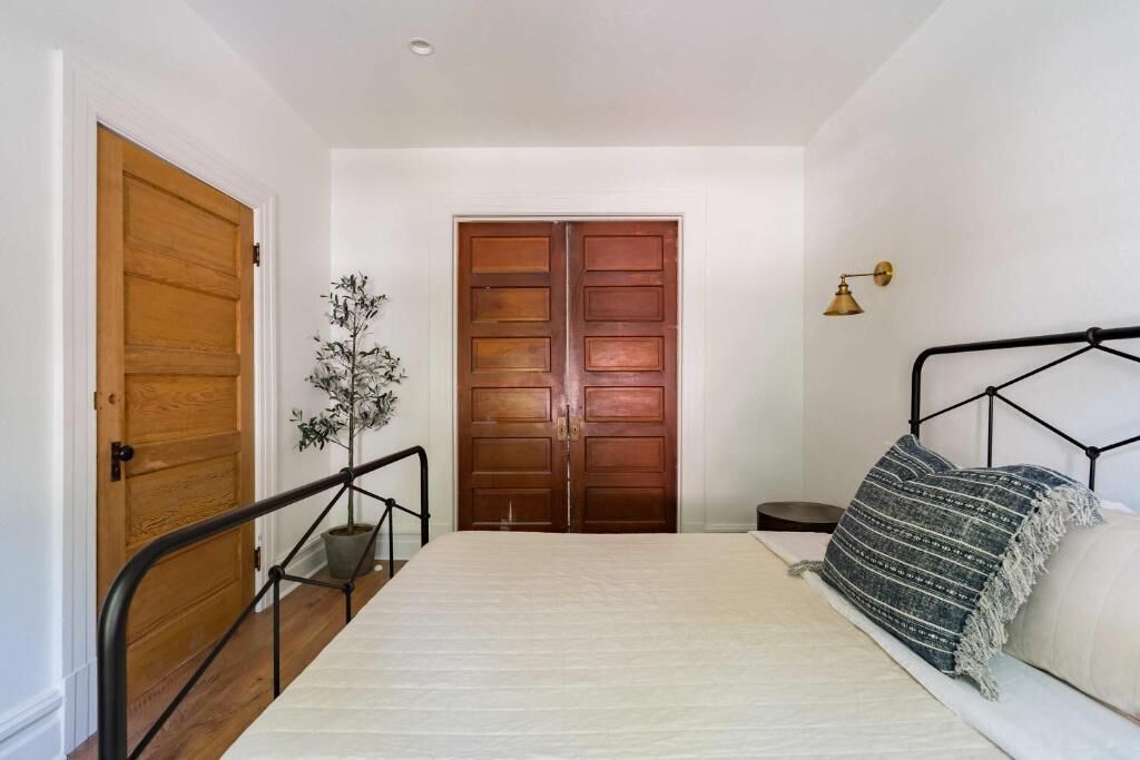 A bedroom with a bed, a wooden door, a small bedside table, a potted plant, and a wall-mounted lamp.