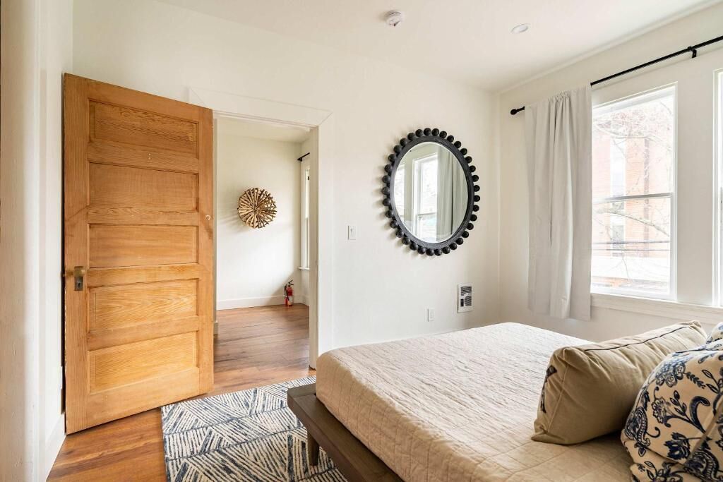 Une chambre avec une porte en bois, un miroir rond, un lit agrémenté d’oreillers à motifs et une vue par la fenêtre.