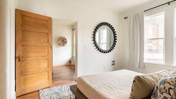 Une chambre avec une porte en bois, un miroir rond, un lit agrémenté d’oreillers à motifs et une vue par la fenêtre.