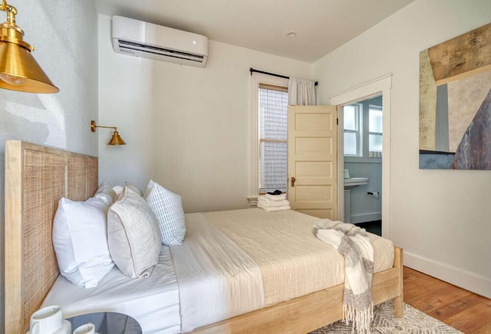 A bedroom with a bed, bedside lamp, air conditioning unit, window with curtains, and a bathroom door.