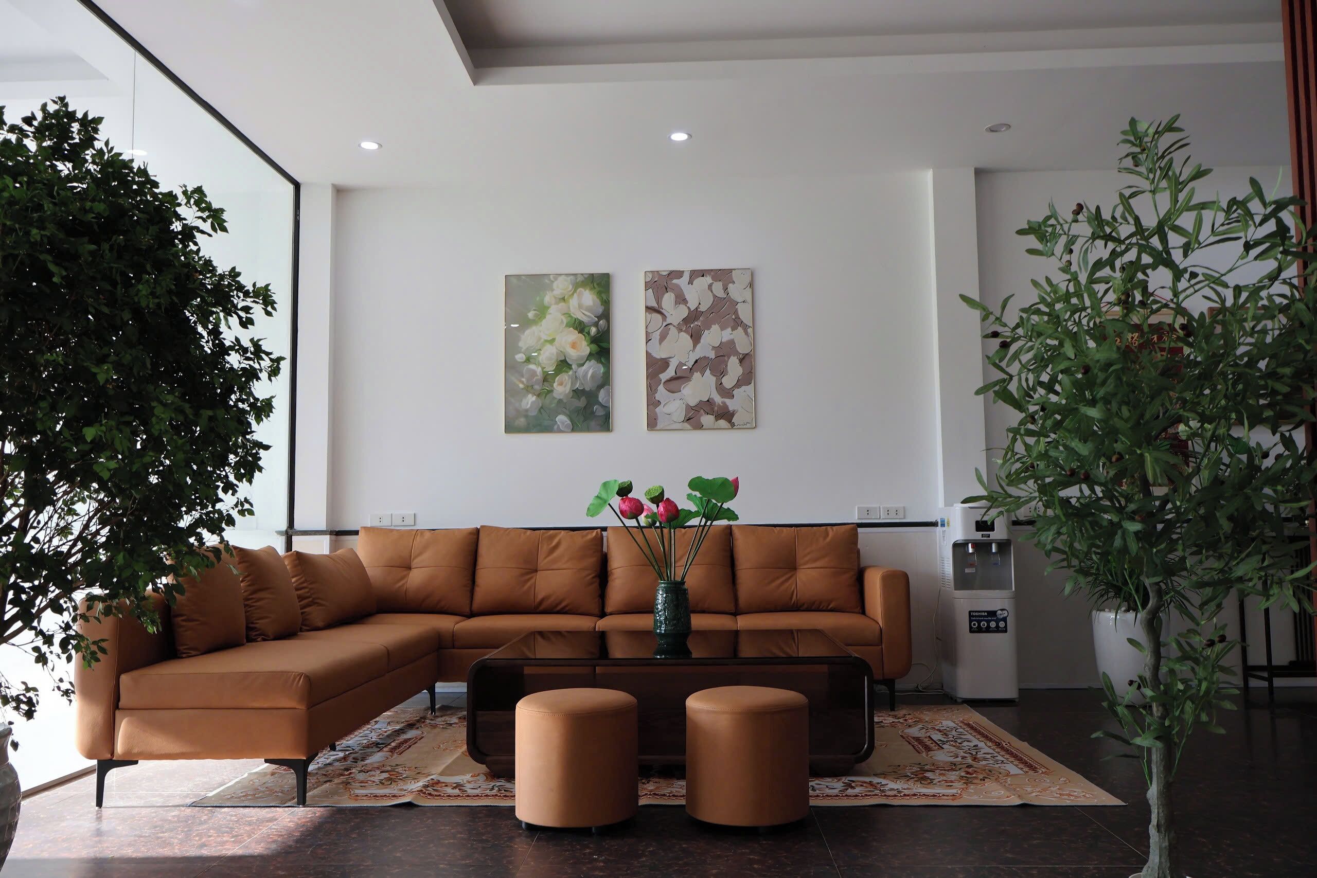 Una sala de estar moderna con un sofá marrón, dos otomanas, una mesa de centro y un jarrón con flores. Hay dos cuadros colgados en la pared, un dispensador de agua y una planta en maceta.