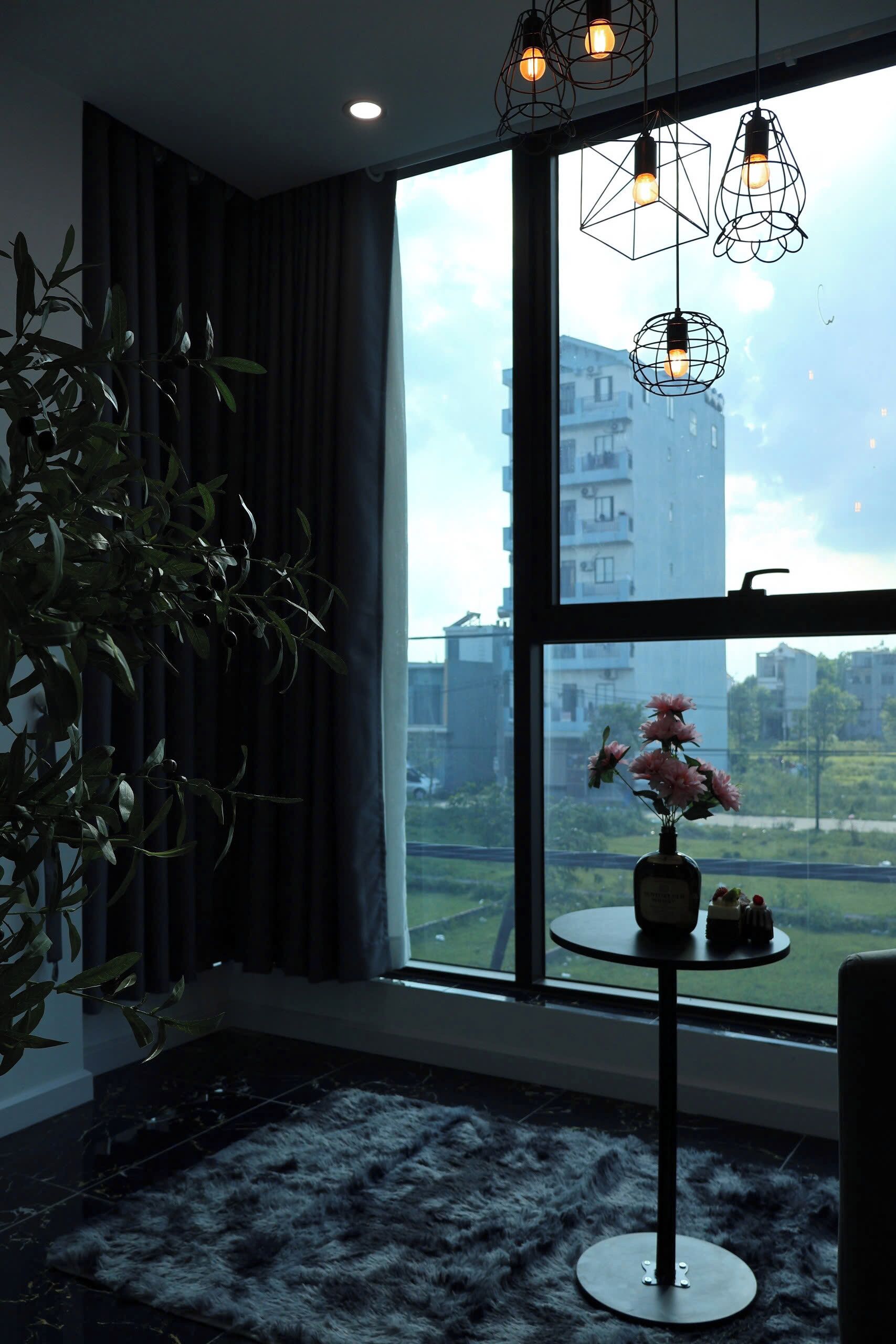 A room with a large window, a potted plant, a round table with a vase, and hanging light fixtures.