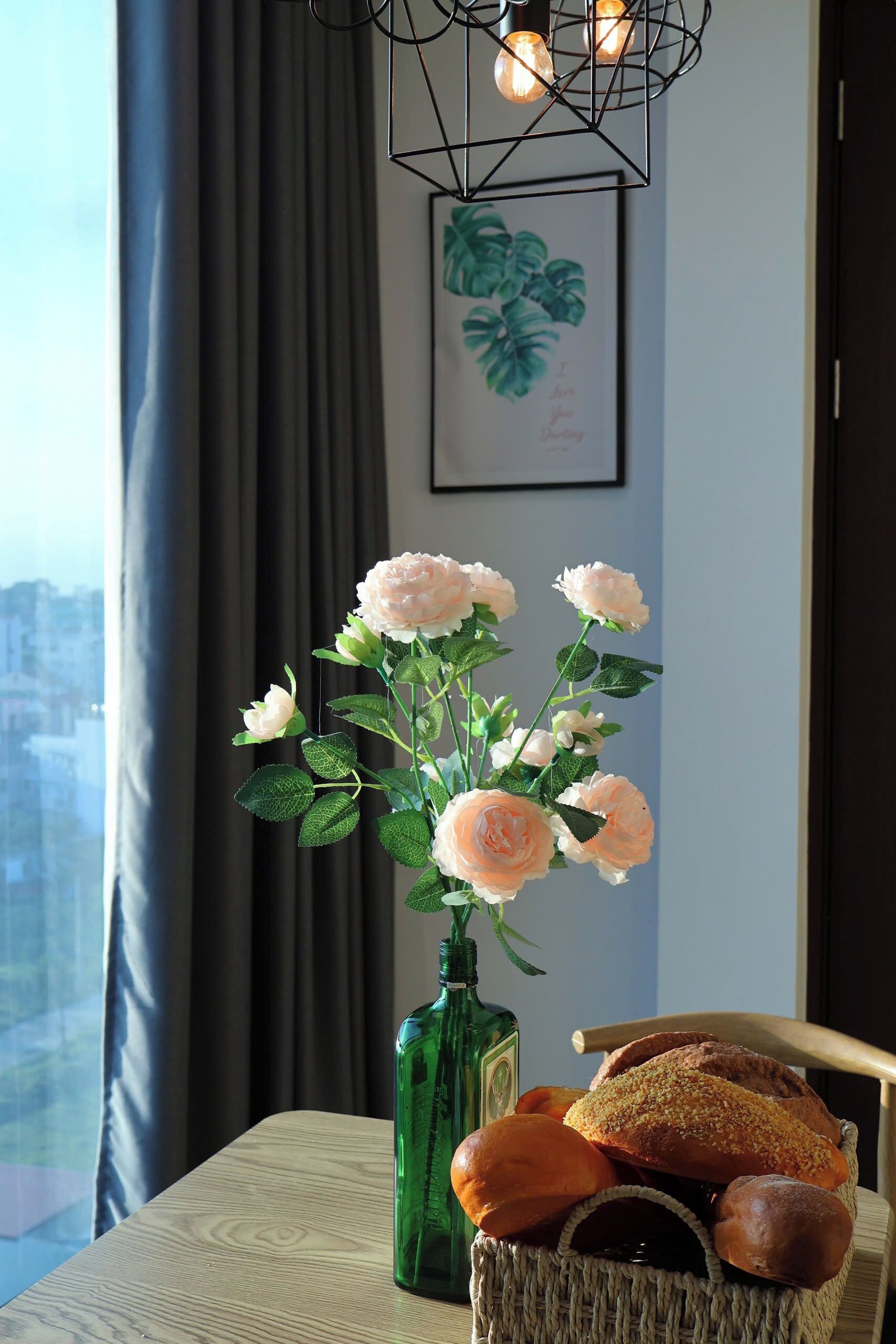 A room with a view of a cityscape, a vase of artificial flowers, and a basket of bread on a table.
