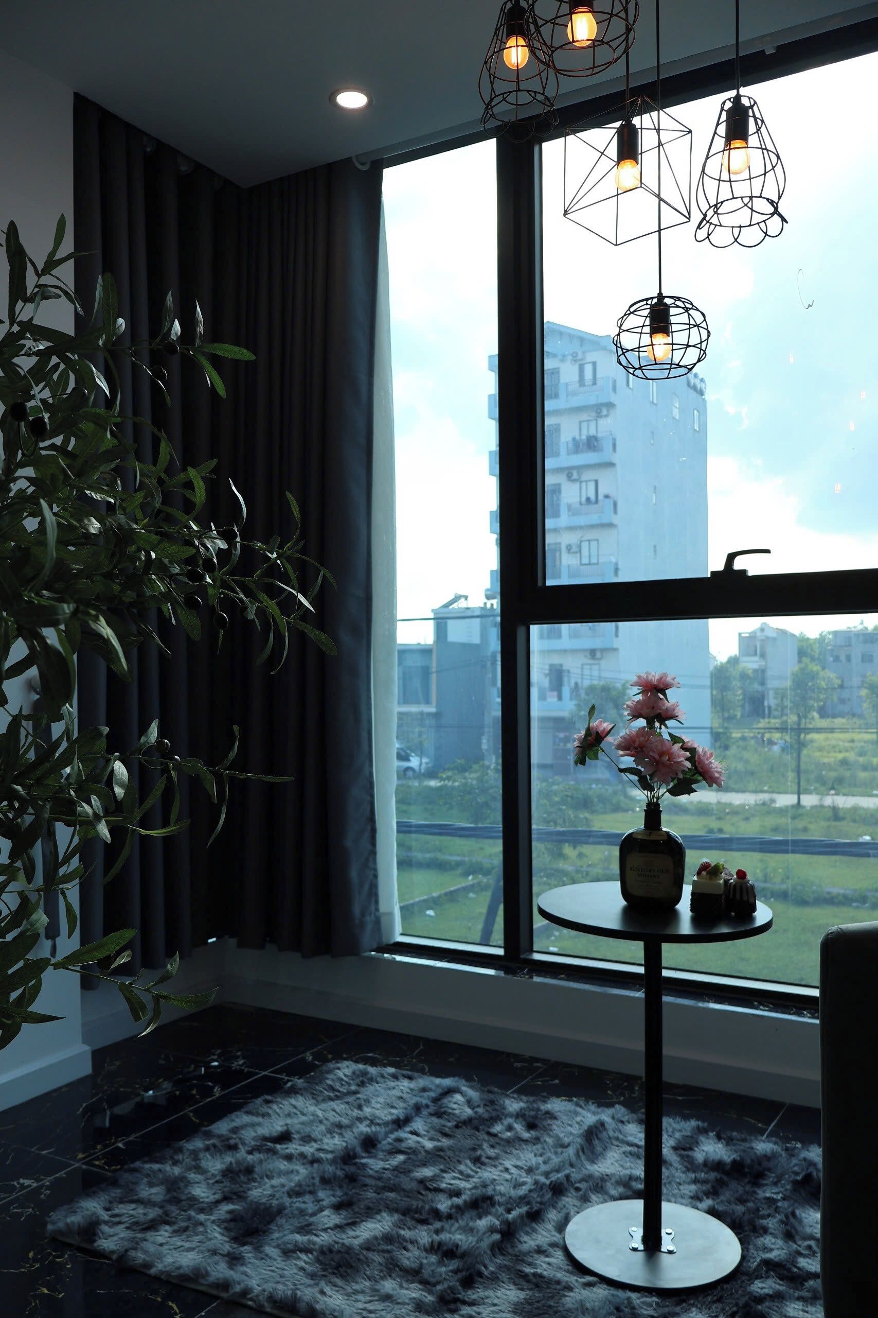 Una habitación con un ventanal grande, una planta en maceta, una mesa redonda con un florero y lámparas colgantes.