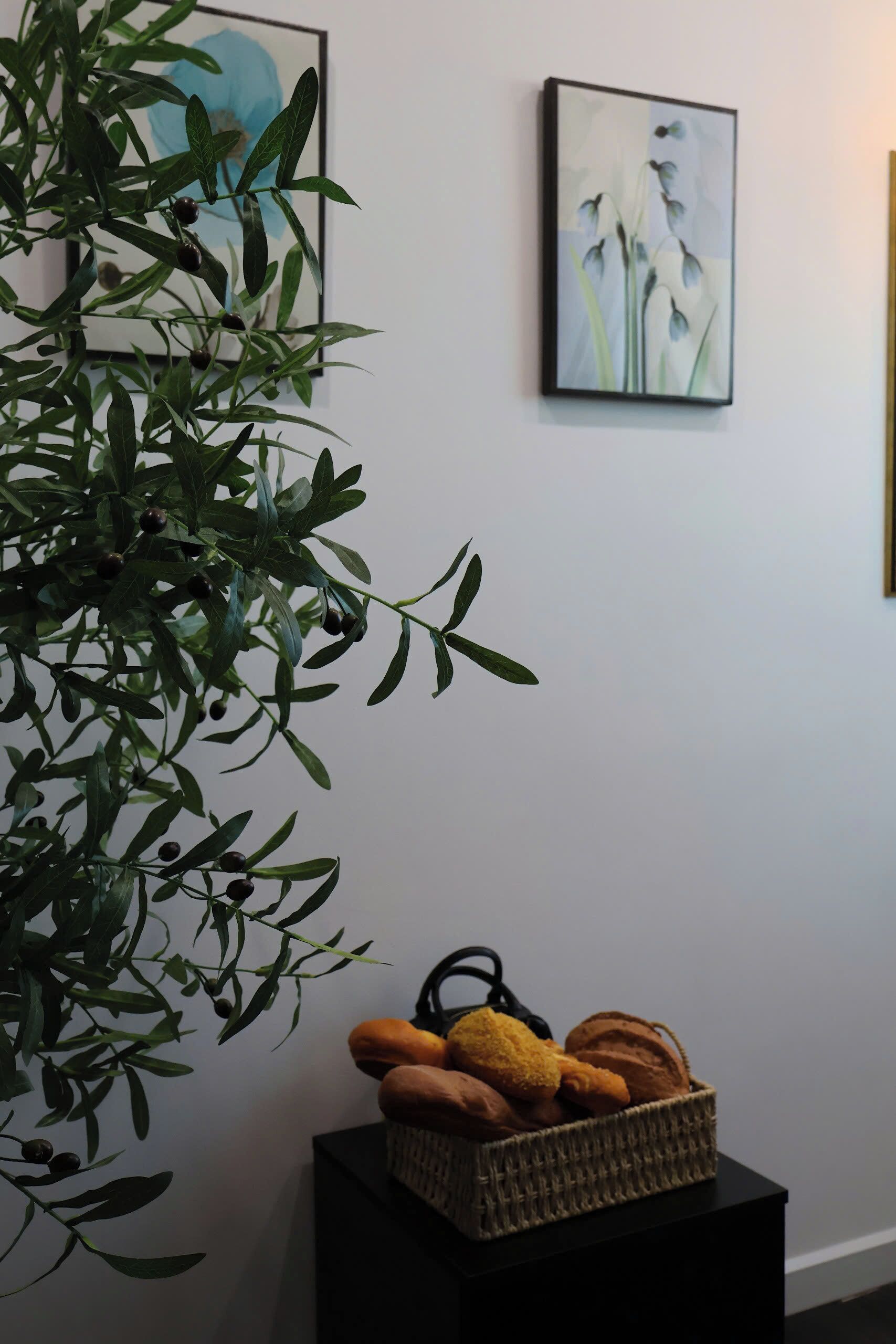 Een kamer met een grote groene plant, twee kunstwerken in lijsten aan de muur en een broodmandje op een zwart bijzettafeltje.