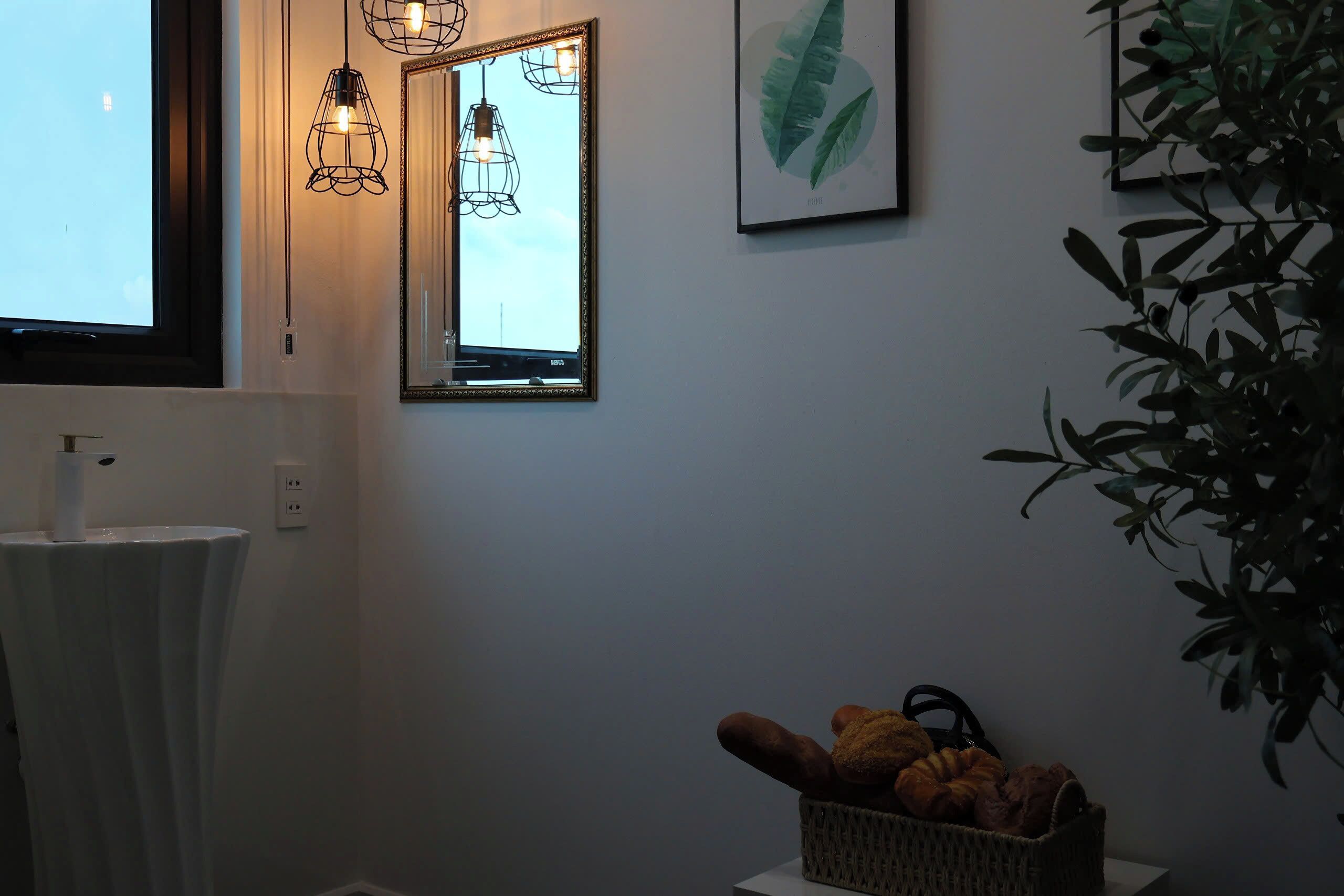 Un bagno con un lavandino bianco, uno specchio, un'opera d'arte con una foglia incorniciata e un cesto di pane su un tavolino.