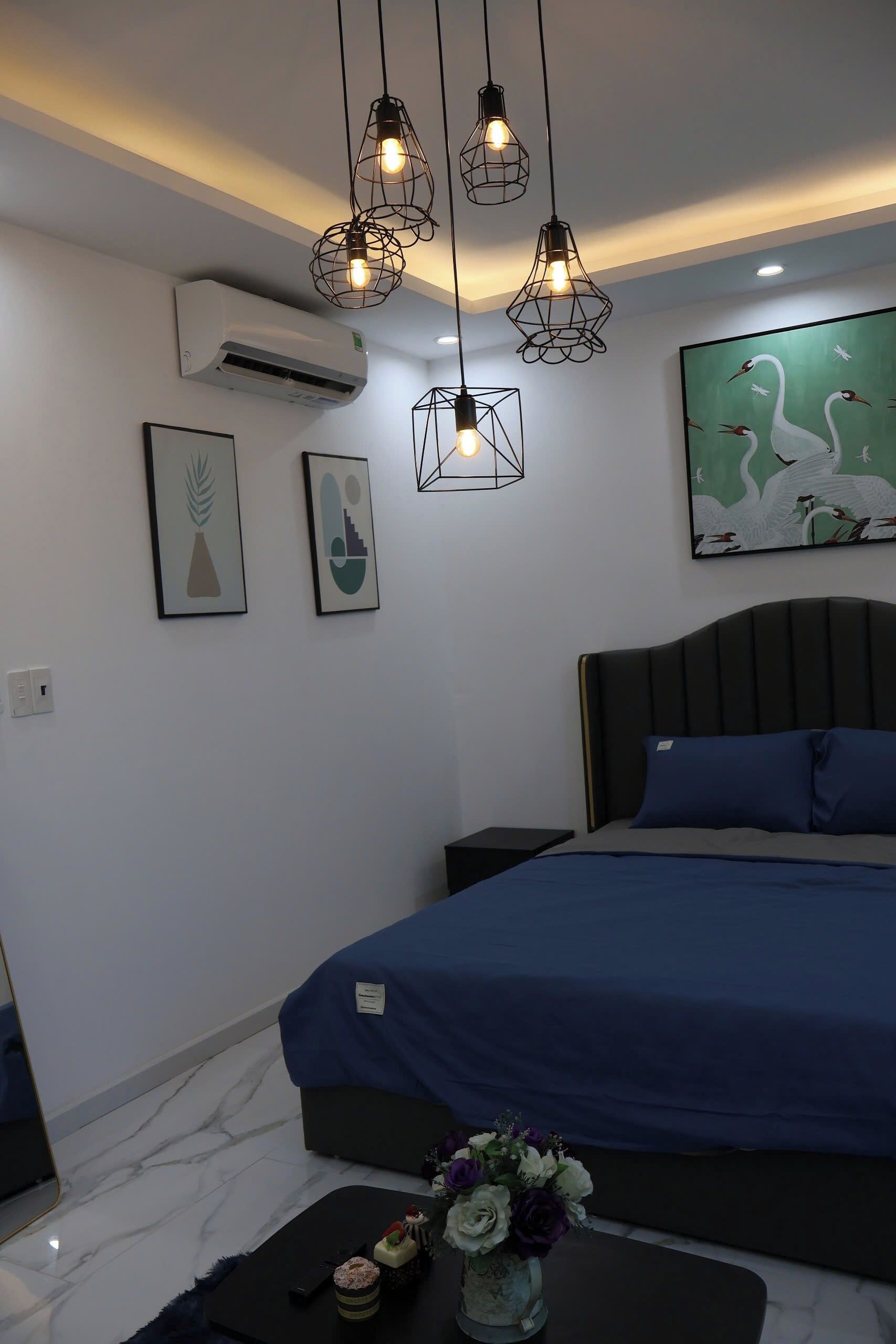 A modern bedroom with a large bed, a bedside table, a nightstand, a wall-mounted air conditioner, and a framed artwork.