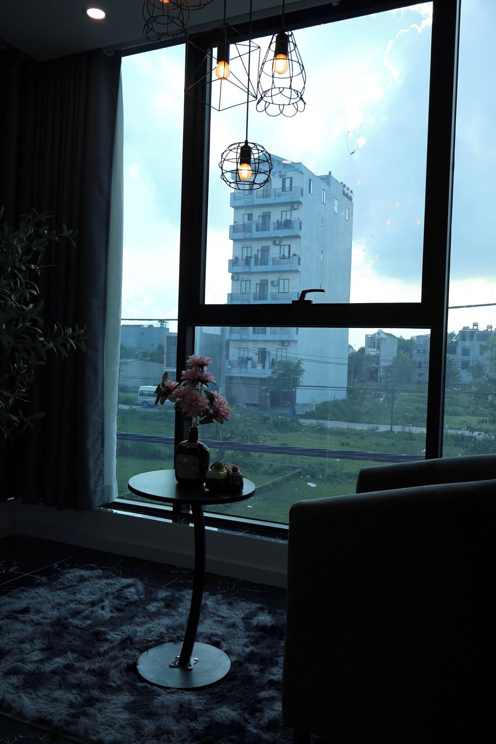 A room with a large window, a potted plant, a vase with flowers, and a table with a bottle and a small object.