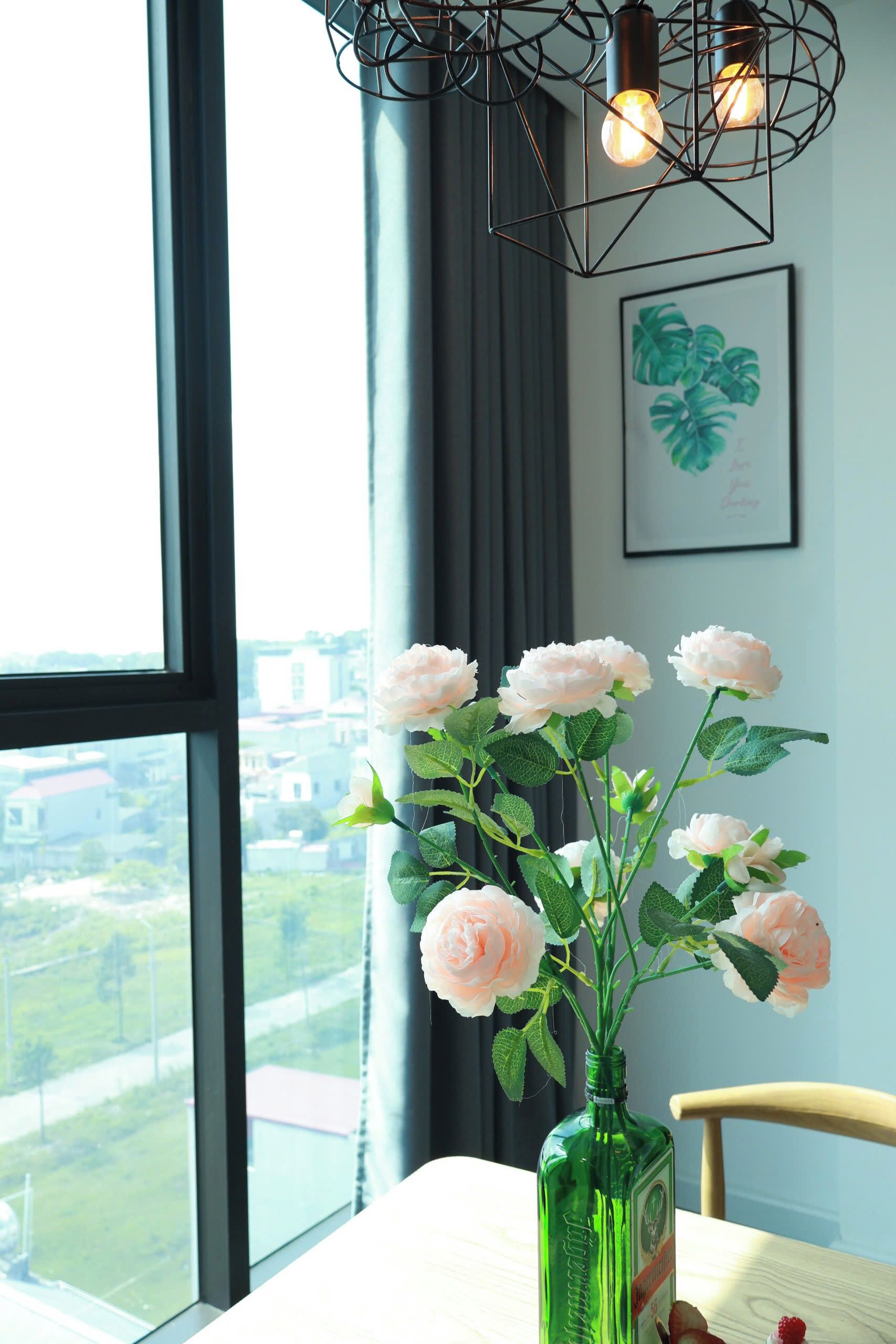 A table with a green bottle vase containing pink roses, a framed plant, and a wooden chair.