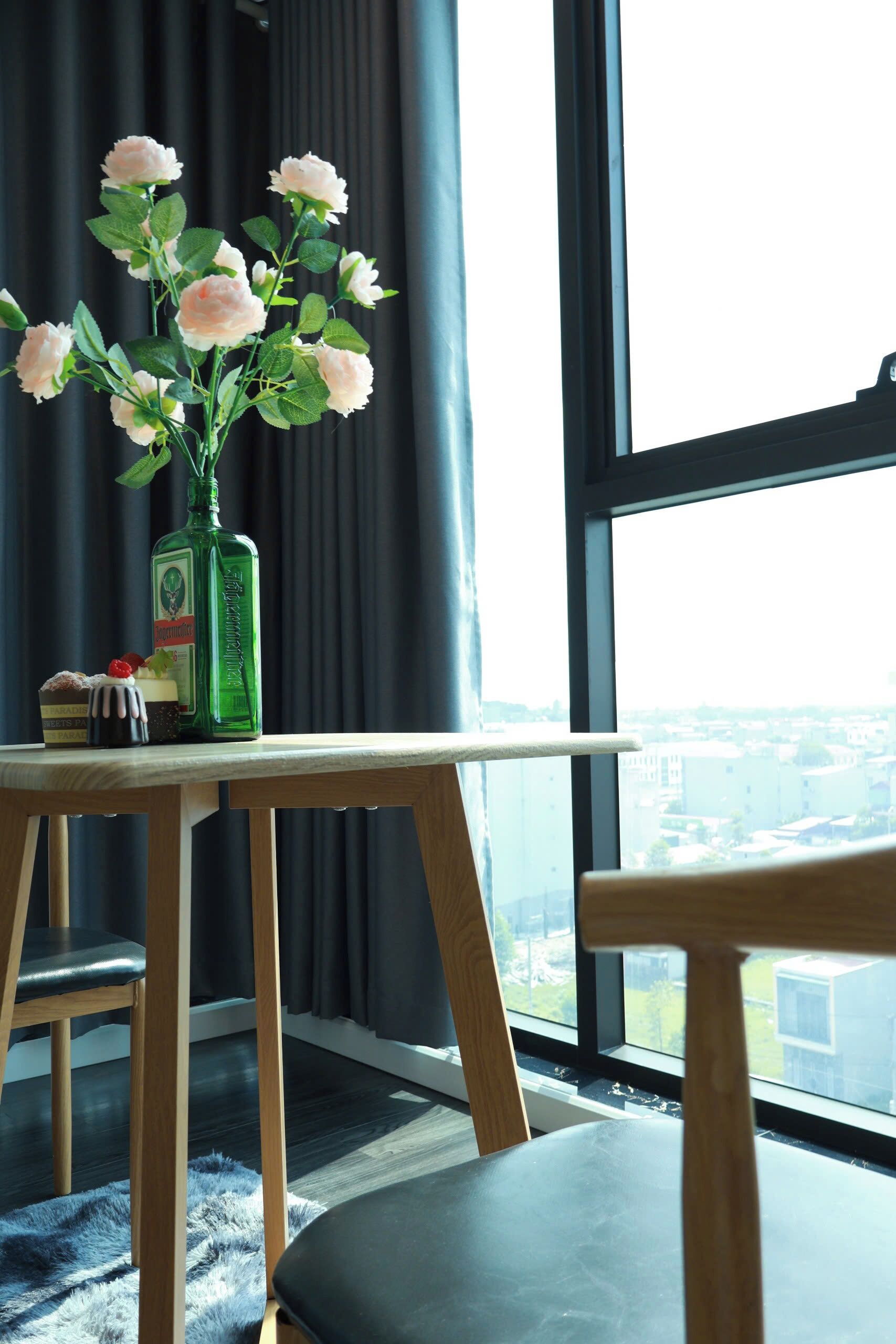 A room with a view of the city through large windows, a wooden table with a vase of flowers, a bottle, and a small cake.