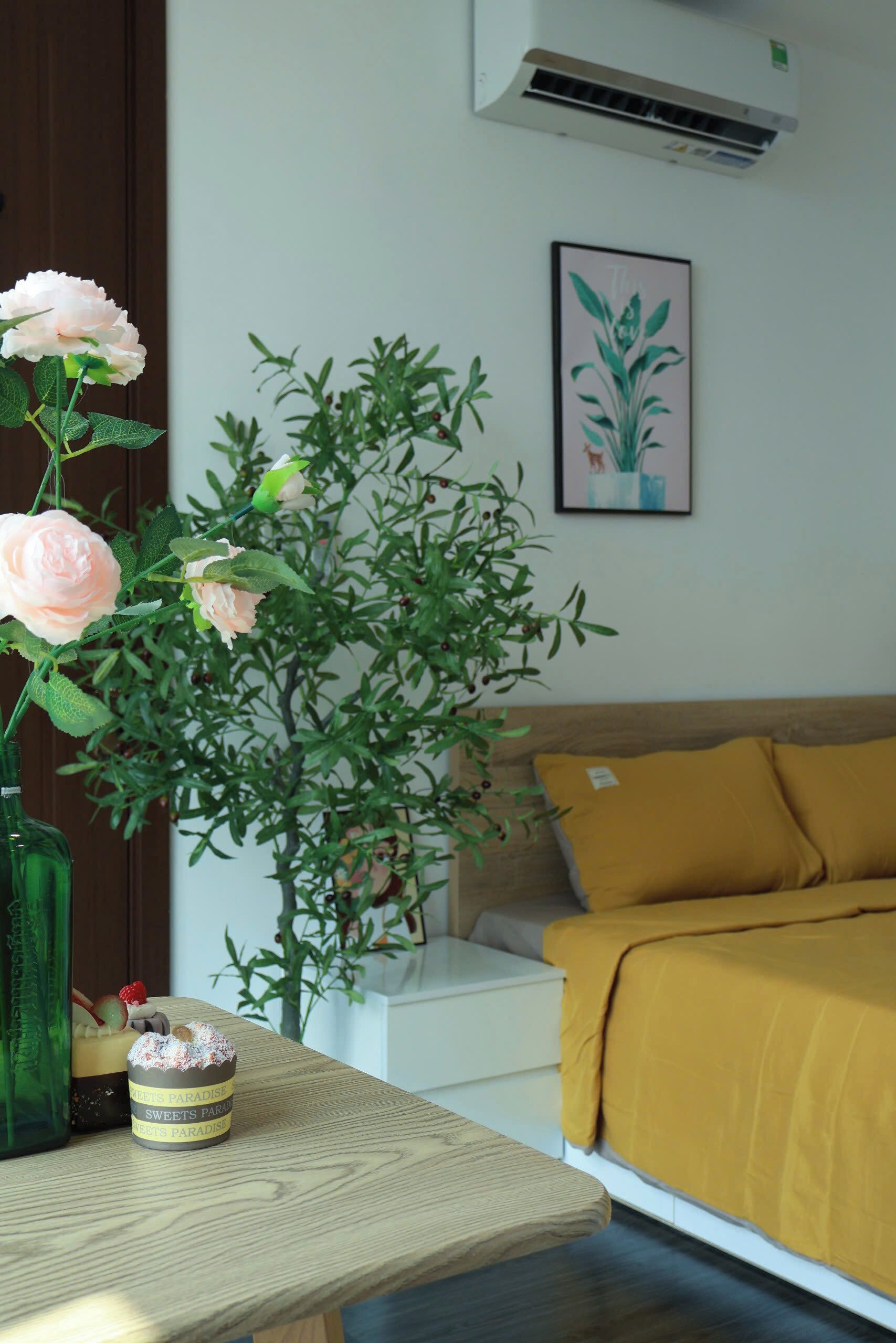 A room with a bed, a bedside table with a vase of flowers, a framed picture on the wall, and an air conditioning unit.