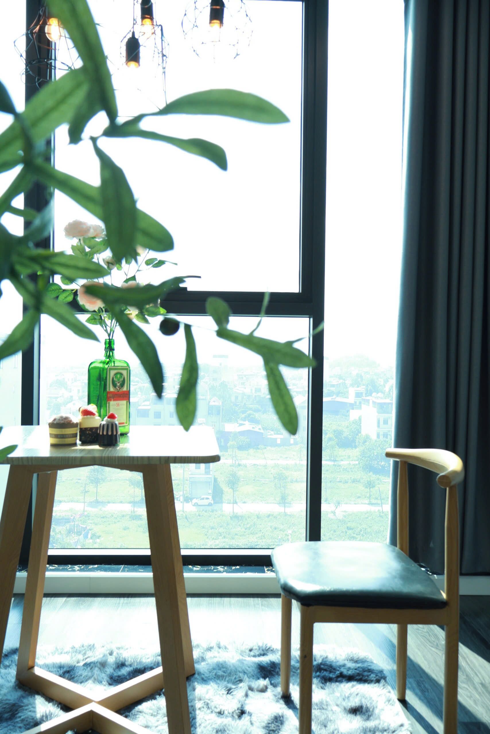 A room with a large window, a wooden chair, a small table with a bottle and two desserts, and a plant in the foreground.