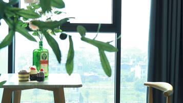 A room with a large window, a wooden chair, a small table with a bottle and two desserts, and a plant in the foreground.