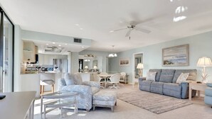 Un salon spacieux comprenant un grand canapé, un canapé plus petit et une table à manger. Il y a également un coin cuisine avec un réfrigérateur et un évier. La pièce est équipée d’un ventilateur de plafond et d’une grande fenêtre qui laisse entrer la lumière naturelle.