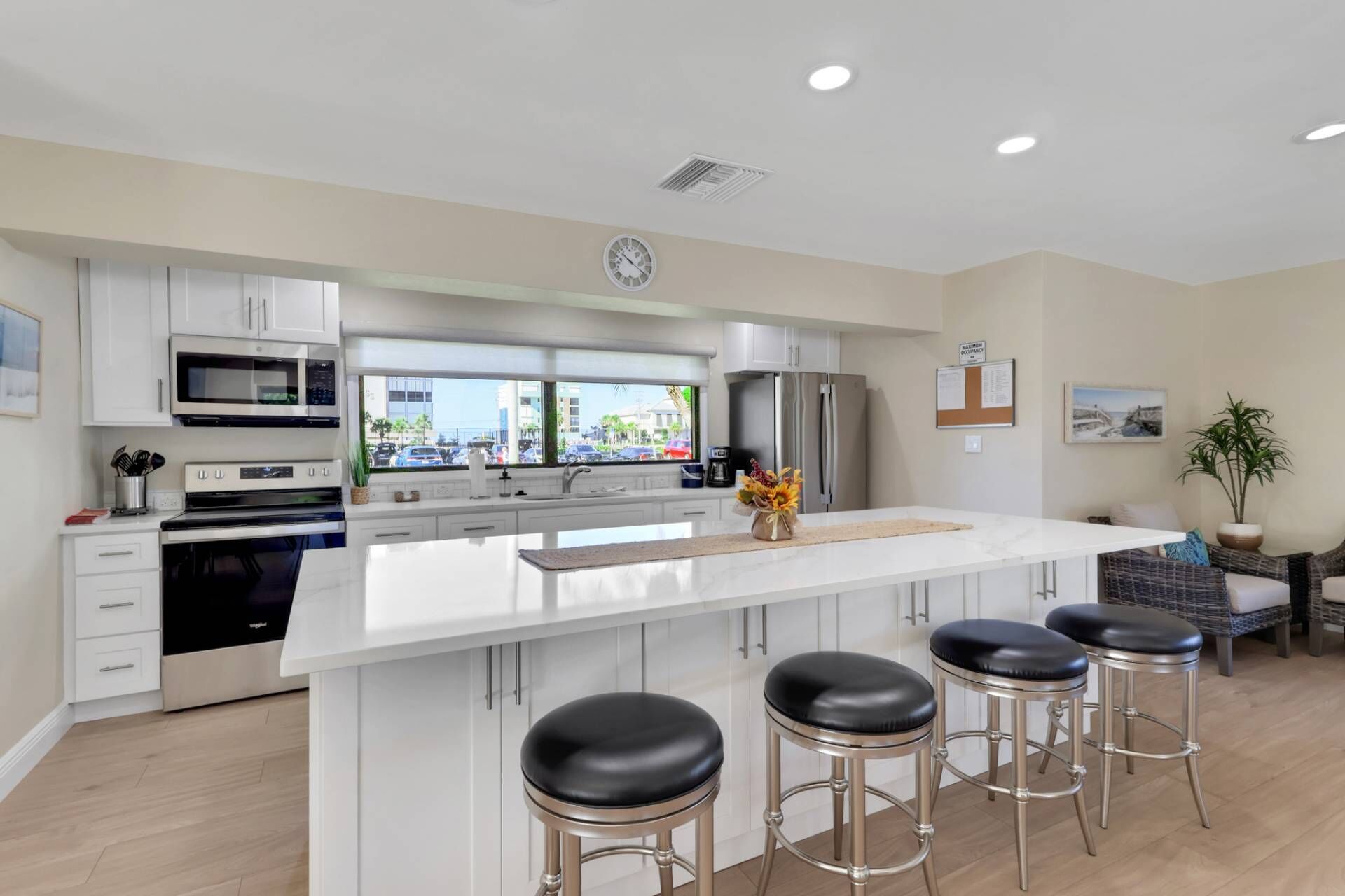 A modern kitchen with white cabinetry, stainless steel appliances, a large island with stools, and a view of the street through the window.