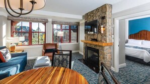 A hotel room with a stone fireplace, a bed, a dining table, and a view of the outdoors through large windows.