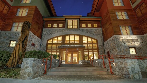 A large, multi-story building with a stone facade and wooden accents, featuring a grand entrance with a glass canopy and a staircase leading up to it.