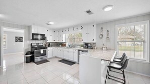 A modern kitchen with white cabinetry, a central island, and a bar area with stools. - Orbital Oasis: Dual Living near NASA (Baytown)