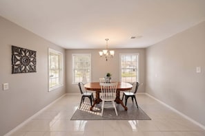 Une salle à manger avec une table ronde, quatre chaises, un lustre et une grande fenêtre avec des stores.