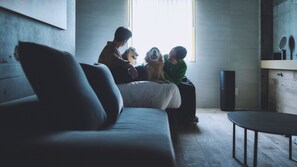 A modern living room with a sofa, a coffee table, and a small round table. There is a window with blinds, a speaker, and a mirror on the wall. - 1HOTEL (Shizuoka)