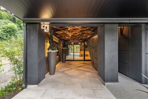 A modern building entrance with a geometric ceiling design, large potted plants, and a pathway leading to a glass door. - 1HOTEL (Shizuoka)