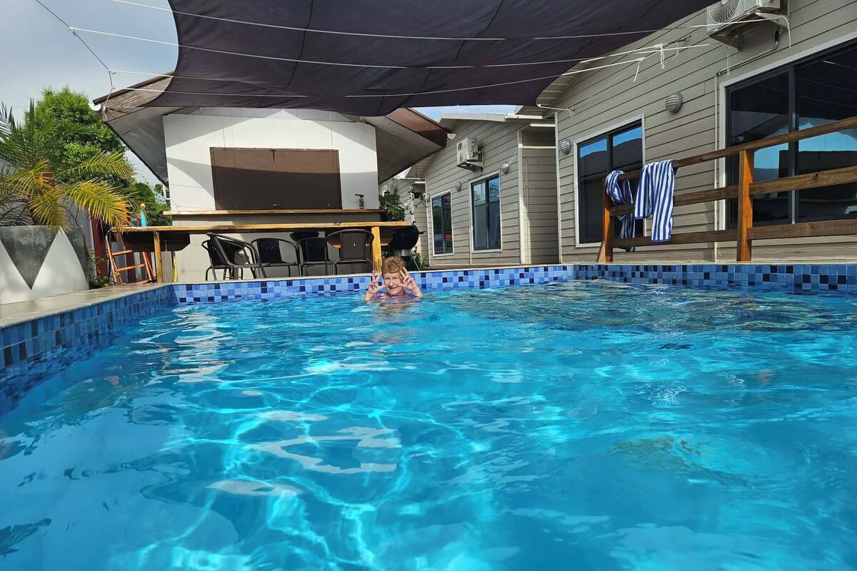 Ein Pool mit gefliestem Rand, eine überdachte Terrasse und ein Haus im Hintergrund.
