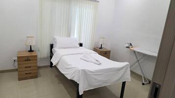 A neatly made bed with white linens, two bedside tables with lamps, a wooden dresser, and a portable ironing board.