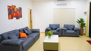 A living room with two grey sofas, a small table with a plant, and a window with blinds. - Sunset Villas (Korotogo)