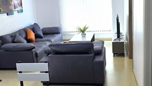 A modern living room with a grey sofa, a white chair, a small table with a plant, and a TV stand. - Sunset Villas (Korotogo)