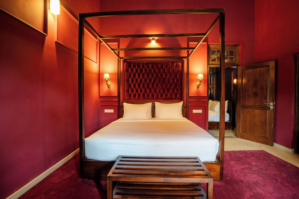 A four-poster bed with a tufted headboard, a wooden bench at the foot of the bed, and two wall-mounted lamps.