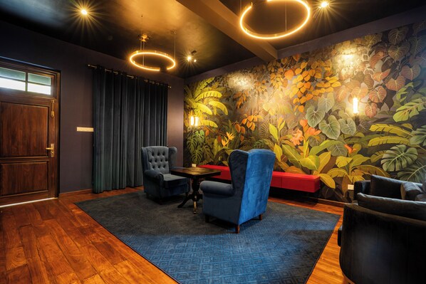A room with a wooden floor, a blue patterned rug, a dark blue armchair, a round table, and a wall mural of tropical leaves. - The Merenchi (Galle)