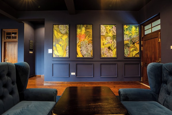 A room with a dark blue wall, a wooden door, and a large window. There are three framed tropical leaf paintings on the wall. - The Merenchi (Galle)