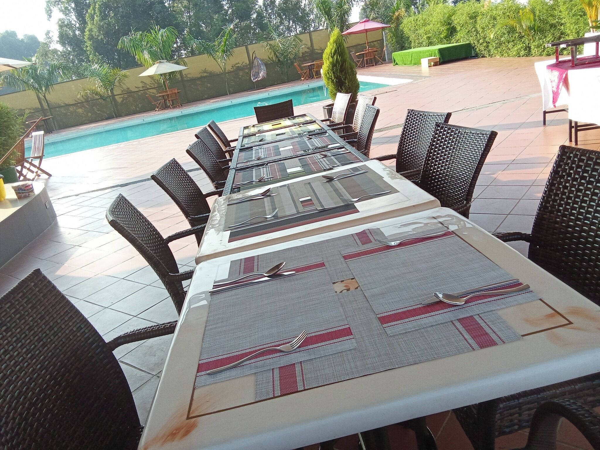A long dining table with black wicker chairs, set with cutlery and placemats, overlooking a swimming pool.
