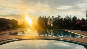 Μια πισίνα με θέα το ηλιοβασίλεμα, που περιβάλλεται από μια πλακόστρωτη βεράντα και δέντρα.