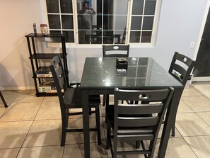 Uma área de refeições com uma mesa de vidro, quatro cadeiras, uma estante de metal preta com jogos de tabuleiro e uma janela com vista para o exterior.