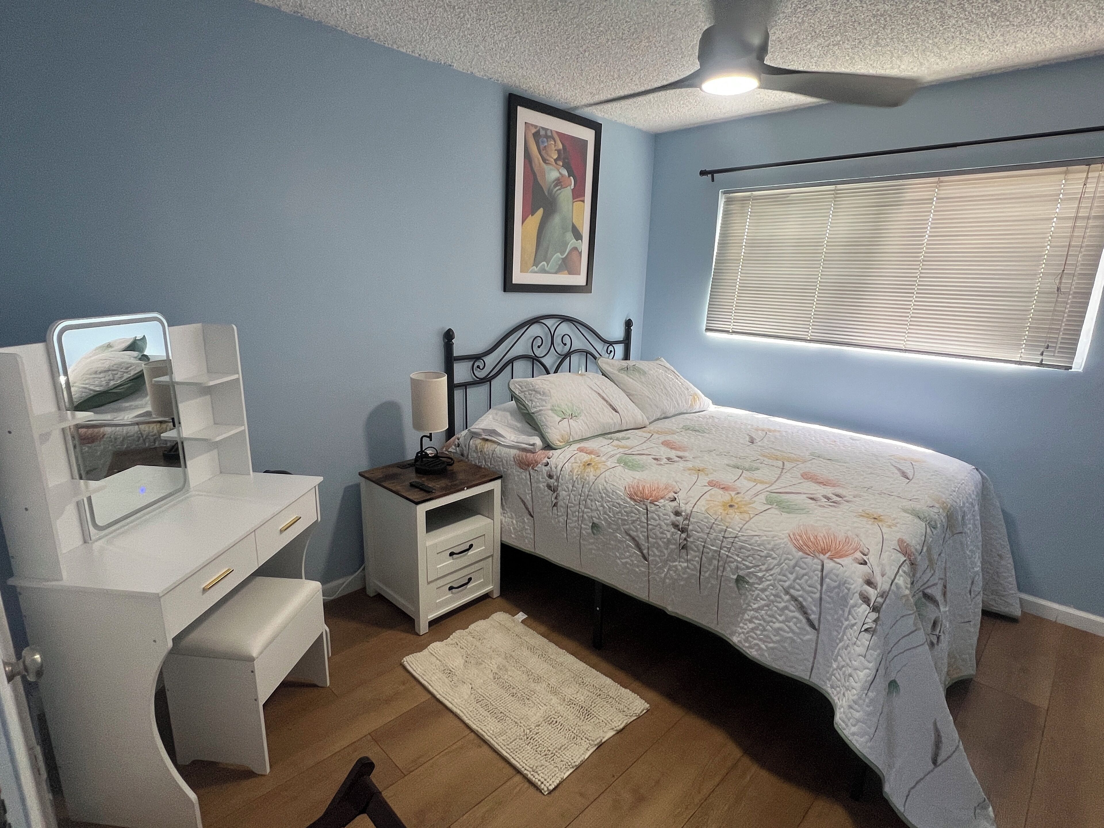 Un dormitorio con cama, escritorio, mesita de noche, espejo y ventana con persianas.