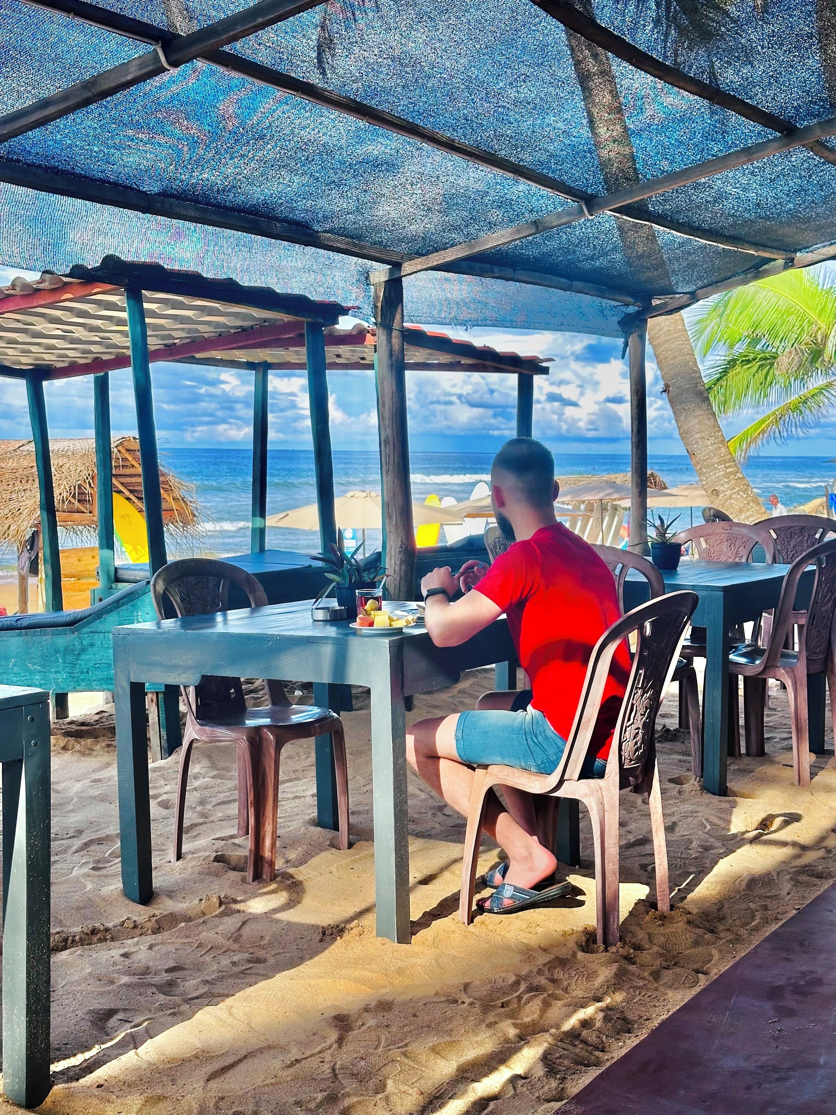 A beachside dining area with tables and chairs under a shaded structure, overlooking the ocean.