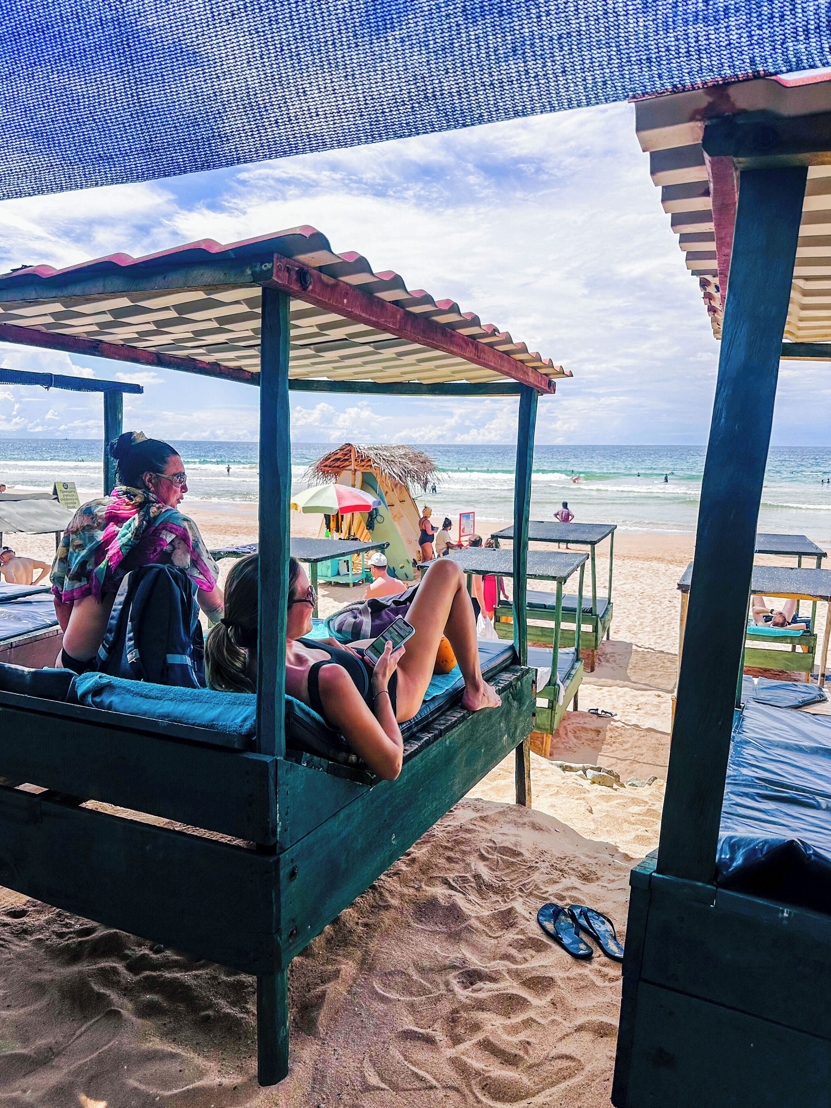 A beach cabana with a thatched roof, a bench, and a table with a person sitting.